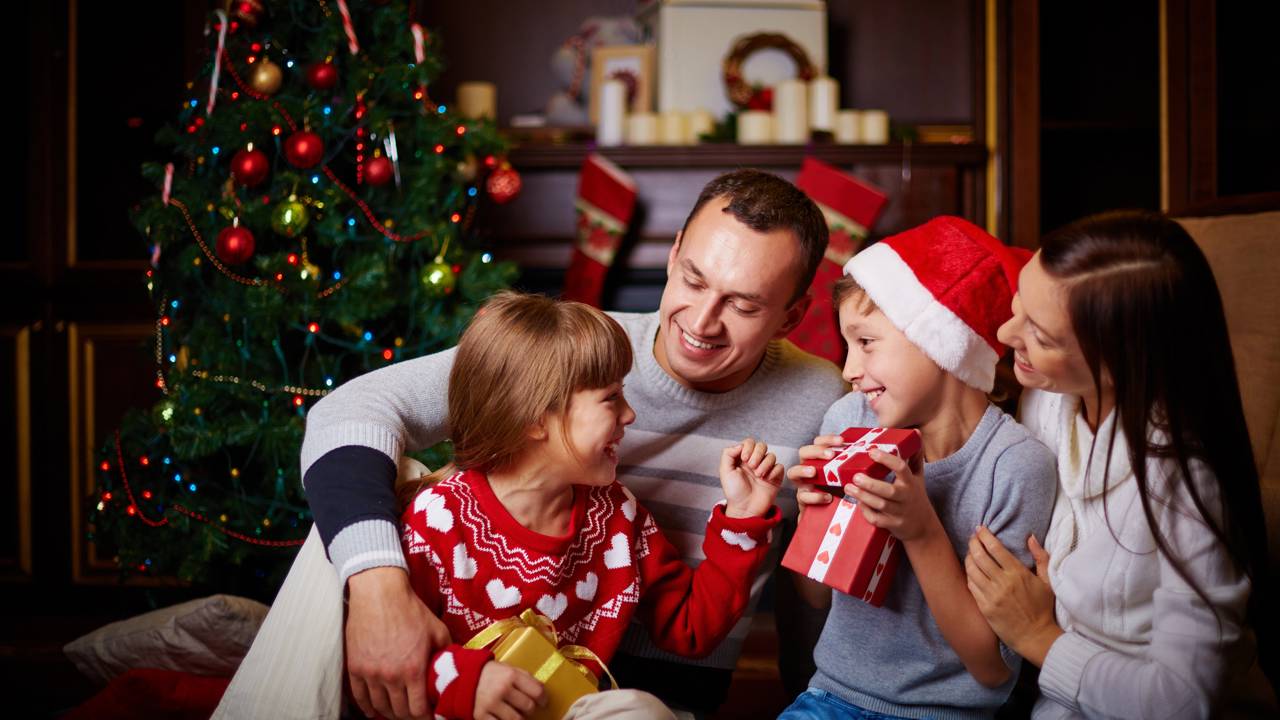 Crianças e adultos brincam com biscoitos, copos e doces de Natal, montando pequenas torres e fazendo desafios simples que reforçam o clima afetivo da celebração.